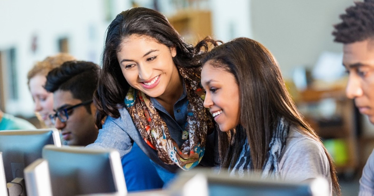 Students sitting in classroom at individual computers. Teacher is standing next to one student pointing something out on her screen. 